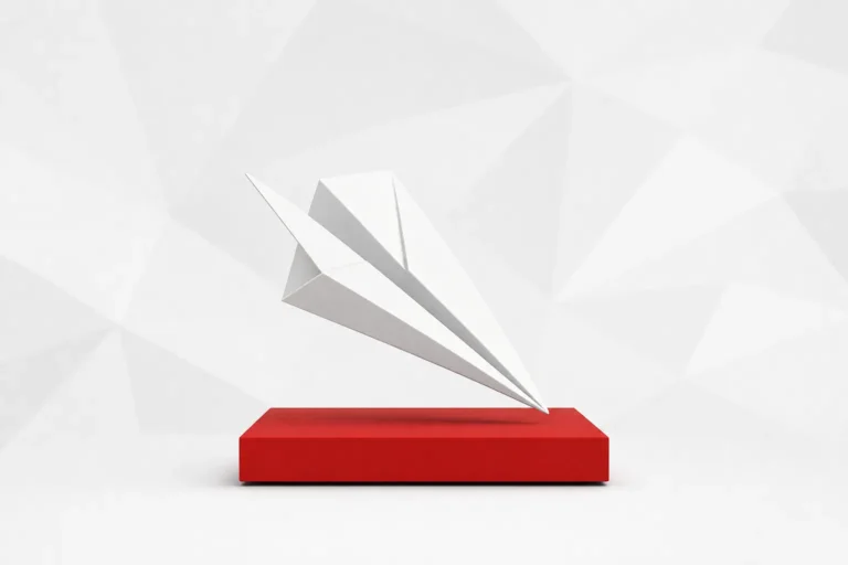Avión de papel blanco que se posa de punta sobre un bloque rojo, ante un fondo gris claro de formas geométricas